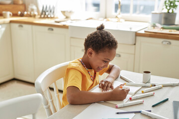 Happy preteen African American girl sitting at table in kitchen enjoying drawing pictures in sketchbook with markers