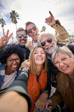 Vertical group diverse cheerful middle-aged tourist friends posing smiling taking photo selfie embracing together with front camera on travel outdoor. Mature happy six people enjoy holidays sunny day 