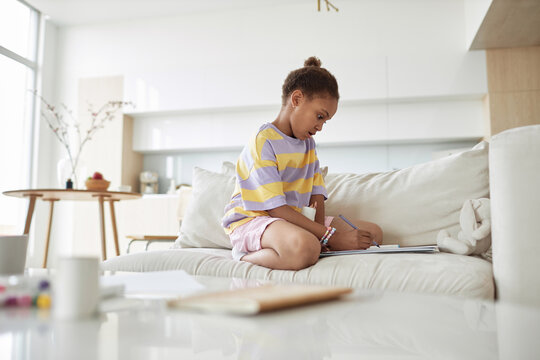Modern black girl sitting on couch at home drawing picture or doing homework using pencil