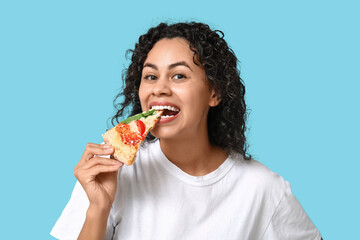 Beautiful young happy African-American woman with slice of delicious pizza on blue background