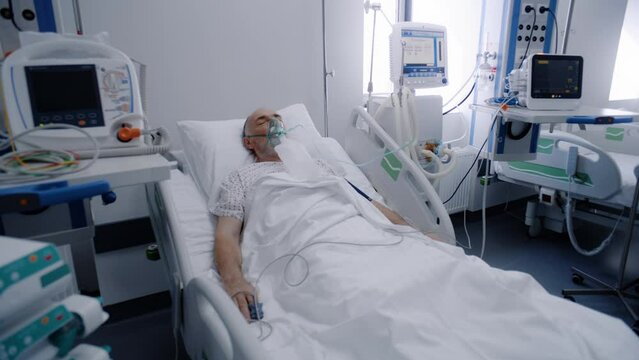 Sick elderly man in oxygen mask sleeps in bed in hospital ward. Old patient during lung ventilation. Equipped emergency room in clinic. Intensive care coronavirus department in modern medical center.
