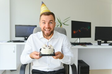 Young happy businessman celebrating his birthday in the office. He holds a cake in his hands.
