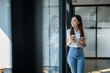 A cheerful young businesswoman engaging in a phone conversation while holding a coffee cup, inside a bright modern office space.