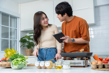 Obraz na płótnie Canvas Happy Family portrait of loving young asian of having fun standing a cheerful preparing food See in the tablet and enjoy cook cooking meat, bread while standing on a kitchen Condo life or home