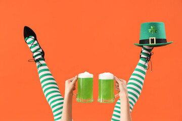 Female legs in green stockings with glasses of beer and leprechaun's hat on orange background. St....
