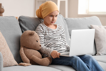 Little girl after chemotherapy with toy bear watching cartoons on laptop at home. International...