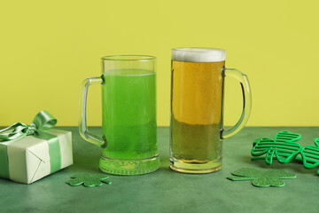 Mugs of beer with plastic eyeglasses, gift box and paper clovers on green grunge table against yellow background. St. Patrick's Day celebration