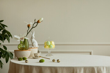 Minimalist composition of easter dining room interior with copy space, gray easter bunny sculpture, colorful eggs, vase with magnolia, beige wall with stucco and personal accessories. Home decor. 