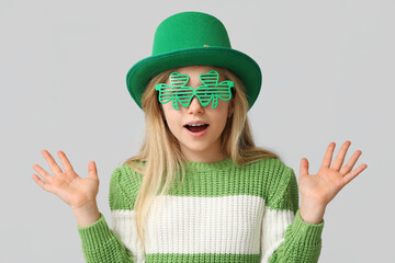 Shocked little girl in leprechaun hat and decorative glasses in shape of clover on grey background....