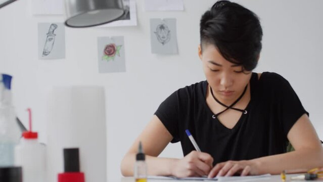 Young concentrated Asian woman sitting at desk in art studio and drawing something with pen