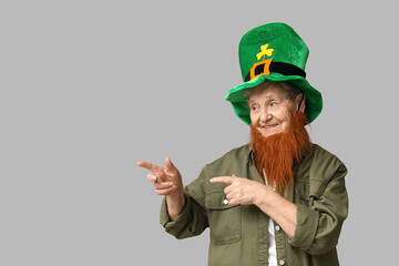 Senior woman in leprechaun hat with red beard pointing at something on grey background. St. Patrick's Day celebration