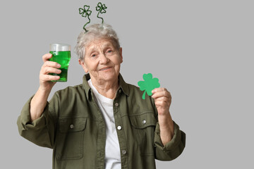Senior woman in headband with glass of beer and clover on grey background. St. Patrick's Day celebration