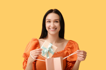 Beautiful young woman with shopping bag and gift box on yellow background. International Women's Day