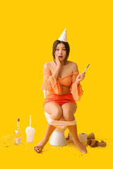 Shocked young Asian woman with pregnancy test sitting on toilet bowl after party against yellow...