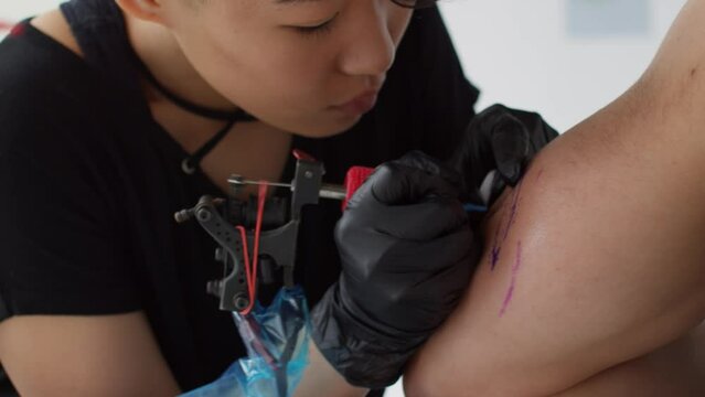 Close up shot of concentrated female tattoo artist using gun for making tattoo on arm of male client in salon