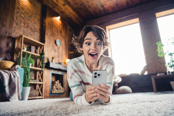 Full size photo of astonished pretty girl laying carpet floor open mouth use smart phone texting daylight flat indoors