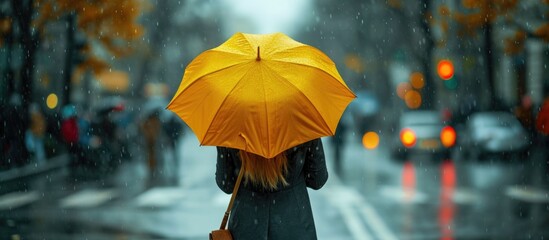 Stylish young woman in a coat with a yellow umbrella walking in the rain on a crossroad.