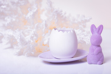 A small purple Easter bunny standing next to an open egg, candles standing on a purple plate, surrounded by boho style decorations on a light background and a white table