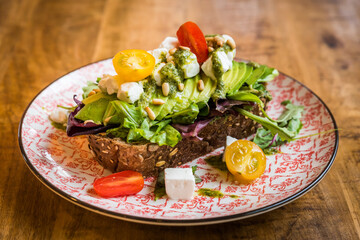 Close up of avocado with feta cheese, tomatoes, pine nuts, pesto and salad on brown bread on a plate on a wooden table
