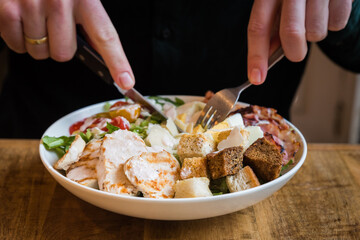 Close up of chicken Caesar salad with bacon, croutons, tomatoes, parmesan cheese, boiled eggs and dressing in a white bowl being eaten with knife and fork on a wooden table