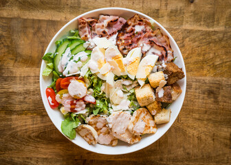Top view of chicken Caesar salad with bacon, croutons, tomatoes, parmesan cheese, boiled eggs and dressing in a white bowl on a wooden table