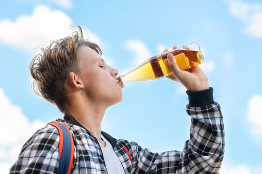 Joyful boy drinking lemonade from a glass bottle against a beautiful sky backdrop. He wears a plaid shirt and a backpack. Vibrant colors