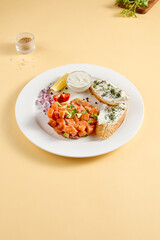 Salmon and avocado tartare with crispy bread and cream cheese, elegantly plated with lemon and dill on a white plate against a yellow background