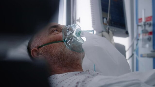Old patient in oxygen mask lies in bed during lung ventilation. Nurse takes care of elderly man, uses monitor. Emergency room with modern equipment in hospital. Intensive care coronavirus department.