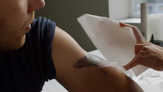 Close up of female tattoo master using paper towel for applying adhesive film dressing on new tattoo of male client