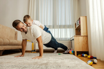 A strong father is doing pushups at home with his son on his backs. - obrazy, fototapety, plakaty