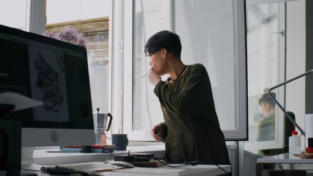 Young Asian woman standing in art studio, looking through open window and smoking a cigarette