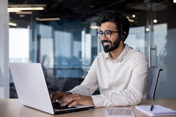 Smiling and confident young Indian man in headset sitting at desk in office and working on laptop