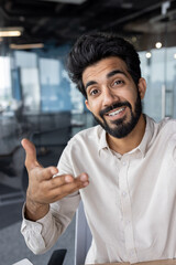 Vertical close-up photo of a young Indian man working in the office and talking to the camera on a video call, smiling and gesturing