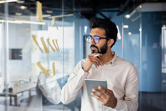 Thoughtful and focused young Indian man standing in office near glass board with tablet and looking at notes. Thinks over new ideas and problem solving - Powered by Adobe