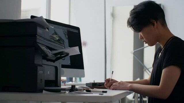 Young Asian woman sitting at table in tattoo salon and drawing sketch on paper from computer screen