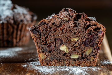 Chocolate muffin with red cherries and walnuts on a wooden table sprinkled with powdered sugar,...