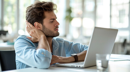 Young man is experiencing discomfort or neck pain while working on a laptop in a bright office setting. - obrazy, fototapety, plakaty