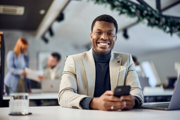 Portrait of a smiling black businessman, looking at the camera, using a mobile phone. - obrazy, fototapety, plakaty