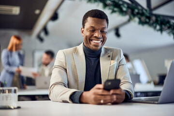 A smiling African man, using a mobile phone, sitting at the office with her colleagues in the background.