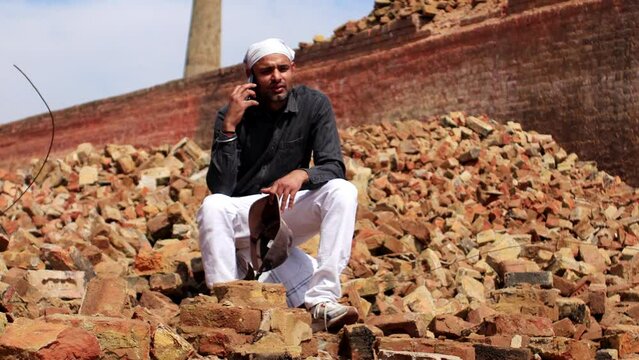 Building contractor sitting on brick stack near brick manufacturing factory and talking on mobile phone