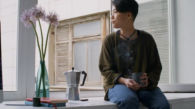 Young Asian woman sitting on window sill in art studio, drinking coffee, smoking a cigarette and then throwing it away