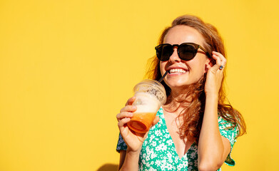 Happy young woman in summer green dress in sunglasses drinking juice and posing in front of yellow...