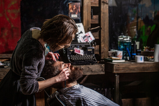 Barber girl shaves his beard with a dangerous razor. Women barber shaves his beard. Barber woman shaving a bearded man in a barber shop