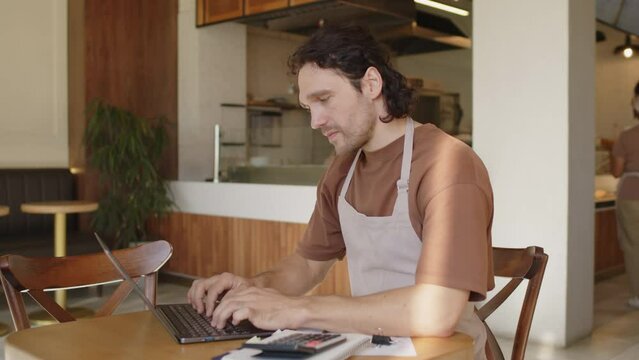 Male bakery owner in apron using laptop and checking receipts while doing accounting during workday in cafe