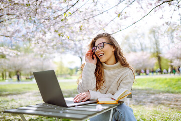 Young curly woman in the blooming  park  with a laptop. Smiling freelancer working outdoors enjoying the good weather. Concept for education, business, blog or freelance.