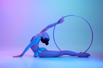 Flexible, talented teen girl, rhythmic gymnast in sparkling bodysuit performing with hoop against gradient studio background in neon light. Concept of sport, grace, competition, art, youth, hobby