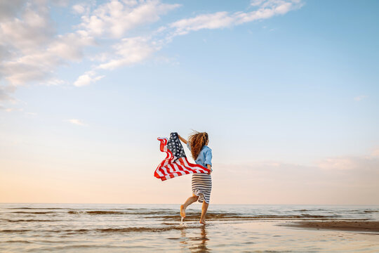 Happy woman holding United States of America flag and running  on the beach at sunset. Patriotic holiday. USA celebrate 4th of July. Independence Day concept