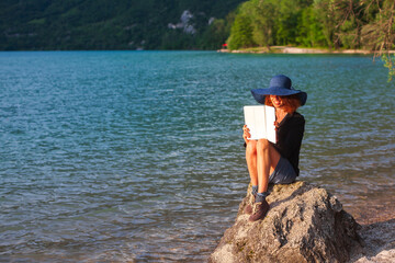 Fototapeta na wymiar Elegant Dressed Woman Using a Digital Tablet While Sitting on a Rock on Coast of a Mountain Lake at Sunset