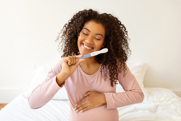 Beaming black expectant mother holding positive pregnancy test and embracing her belly