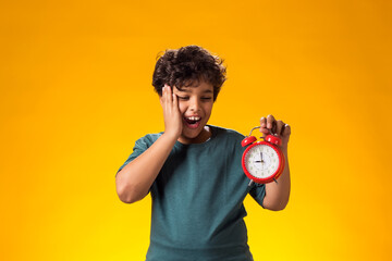 Surprised child boy holding alarm clock over yellow background. Time management concept